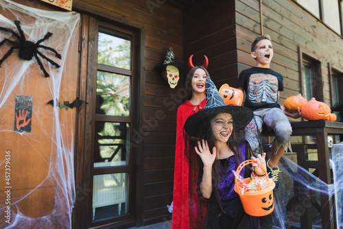 girl in witch hat waving hand near excited kids in spooky halloween costumes on porch with decoration