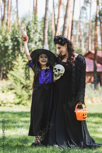 excited girl in witch halloween costume holding toy hand and skull near mom