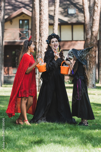 astonished woman with daughters in halloween costumes holding buckets with candies in forest near blurred cottage