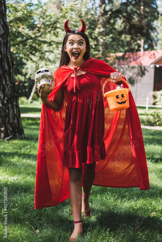 astonished girl in demon halloween costume shouting while standing with skull and bucket of candies outdoors