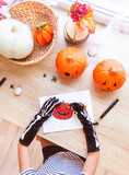 A child in black gloves with bones draws a laughing pumpkin while sitting by the window, surrounded by pumpkins and spiders, top view. Preparing for the holiday, decorating the house for Halloween.