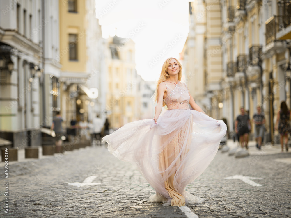 Obraz premium Pretty smiling young woman with long blond hair in elegant flying light dress running along the street