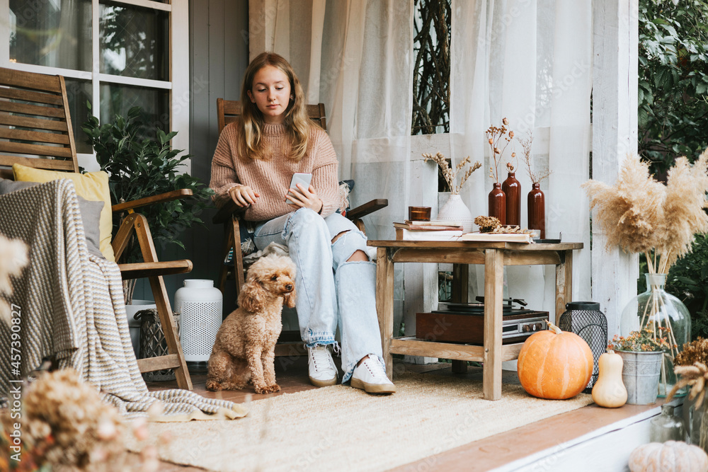 beautiful teenage girl in cozy warm knitted sweater sits and takes selfie on phone with poodle dog on porch of backyard decorated with pumpkins and dry grass, cozy and comfortable autumn lifestyle beautiful teenage girl in cozy warm knitted sweater sits and takes selfie on phone with poodle dog on porch of backyard decorated with pumpkins and dry grass, cozy and comfortable autumn lifestyle