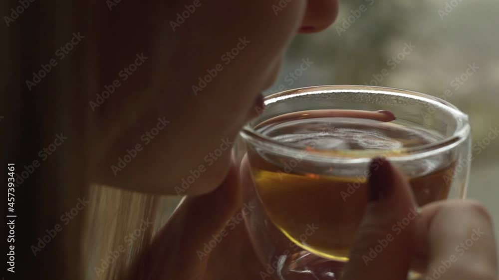 A young girl is sitting near the window and holding a glass mug with warm tea blowing cool air into her cup A young girl is sitting near the window and holding a glass mug with warm tea blowing cool air into her cup