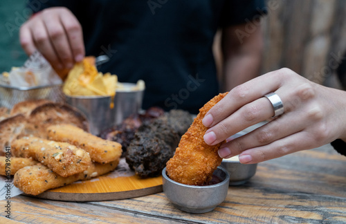 Finger food. Couple sharing a dish with fried mozzarella and chicken sticks, nachos with cheddar and chard fritters. Closeup of a young woman hand dipping a fried chicken stick in hot sauce. 