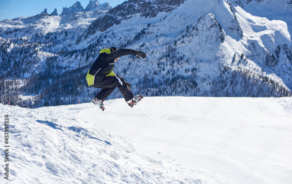 Chłopiec w stroju narciarskim wykonujący skok ze śnieżnej góry. Zimowy krajobraz górski, w tle skaliste, ośnieżone szczyty górskie. - obrazy, fototapety, plakaty 