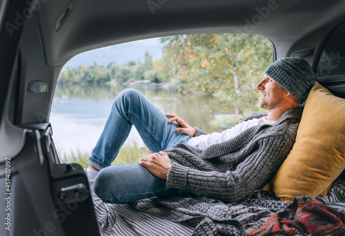 Smiling middle-aged man dressed in warm knitted clothes and jeans lying in the cozy car trunk and enjoying the mountain lake view. Warm early autumn auto traveling concept image. photo