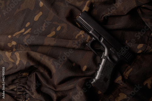 Black airsoft pistol lies against the background of a soldier in dark camouflage. A black pistol is lying on a camouflage soldier's jacket. A firearm that makes you think about self-defense. Top view