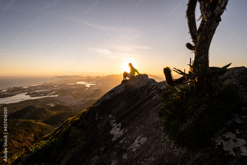 Man sitting on rocky mountain edge next to big cactus on sunset Man sitting on rocky mountain edge next to big cactus on sunset