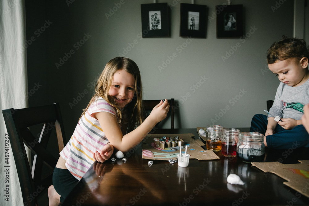 Girl smiling while doing art project at the table with her siblings Girl smiling while doing art project at the table with her siblings