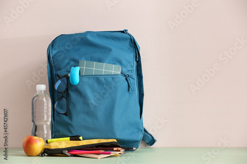 Stylish school backpack with stationery, eyeglasses, bottle of water and apple on table