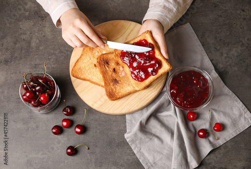 Woman spreading sweet cherry jam onto toast on dark background