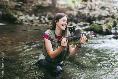 Fototapeta Naklejka Na Ścianę i Meble -  A young woman, who does sports fishing, fishes on a fast mountain river and holds a trout she caught in her hand.
