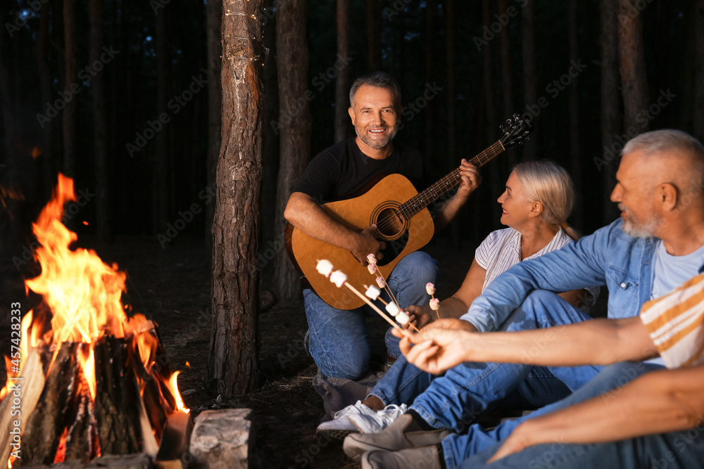 Mature people with marshmallows at barbecue party near fire on summer evening