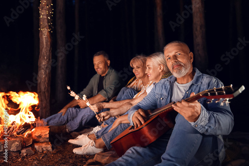 Mature man with guitar near fire at barbecue party on summer evening