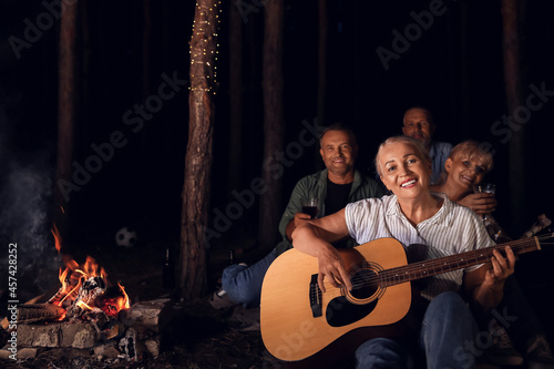 Mature woman playing guitar near fire at barbecue party on summer evening