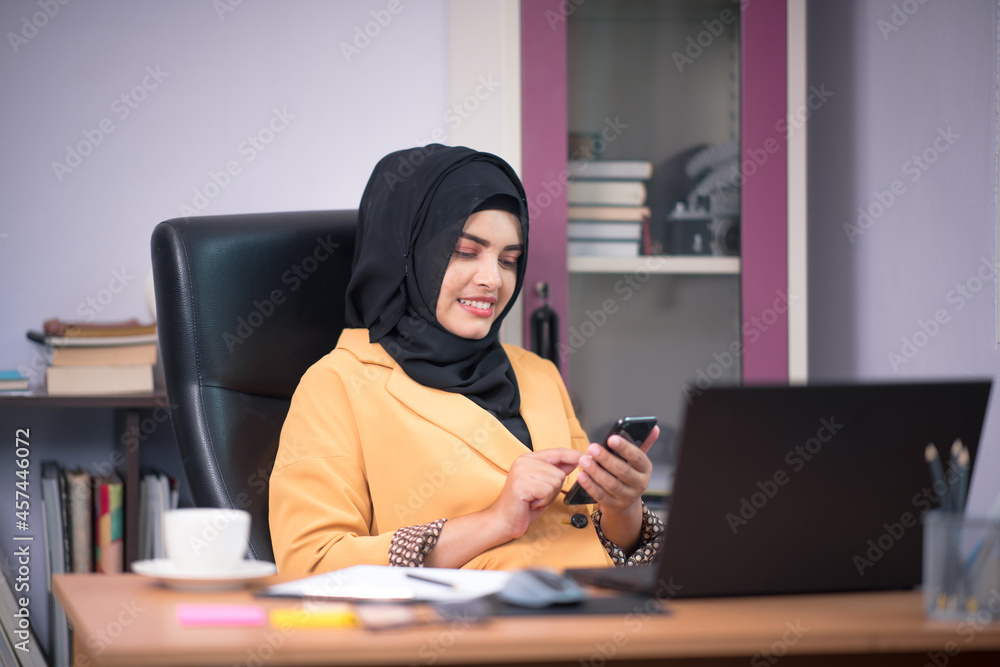 An office worker for an Asian Muslim woman sitting in front of a laptop computer at her desk and talking on a mobile phone and working at the office. An office worker for an Asian Muslim woman sitting in front of a laptop computer at her desk and talking on a mobile phone and working at the office.
