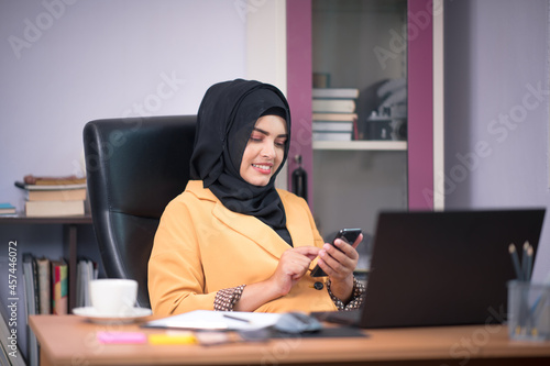 An office worker for an Asian Muslim woman sitting in front of a laptop computer at her desk and talking on a mobile phone and working at the office.