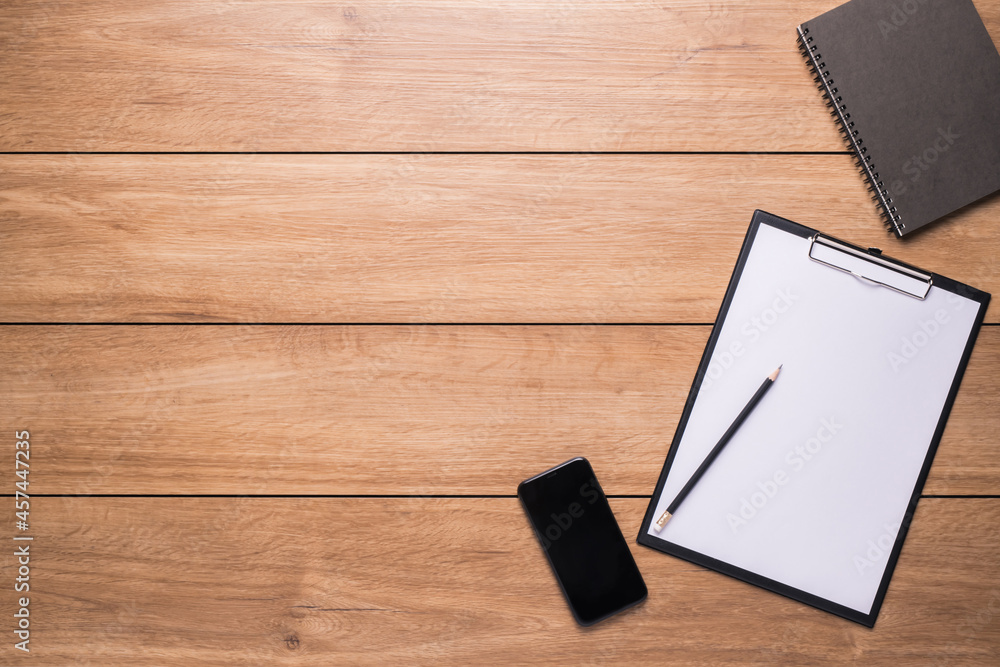 A clipboard with note-taking paper on a pencil and a mobile phone were placed on a wooden desk. A clipboard with note-taking paper on a pencil and a mobile phone were placed on a wooden desk.
