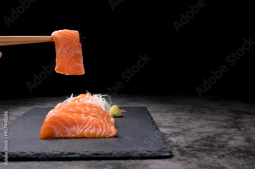 The hands were holding the chopsticks to hold the salmon sashimi, which was arranged on a black stone plate on an old table, with copy space.
