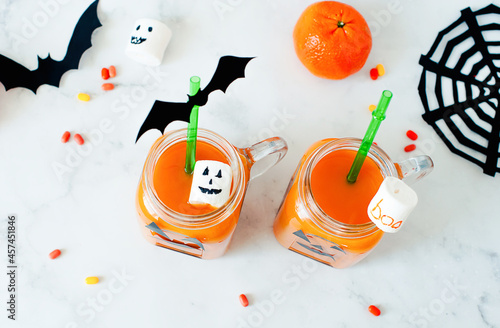 Drink for Halloween. hildren's and party of Halloween treats. Pumpkin orange cocktail in glasses, decorated with creepy faces scary marshmallows, bats, cobwebs on a white marble table. Copy space. photo