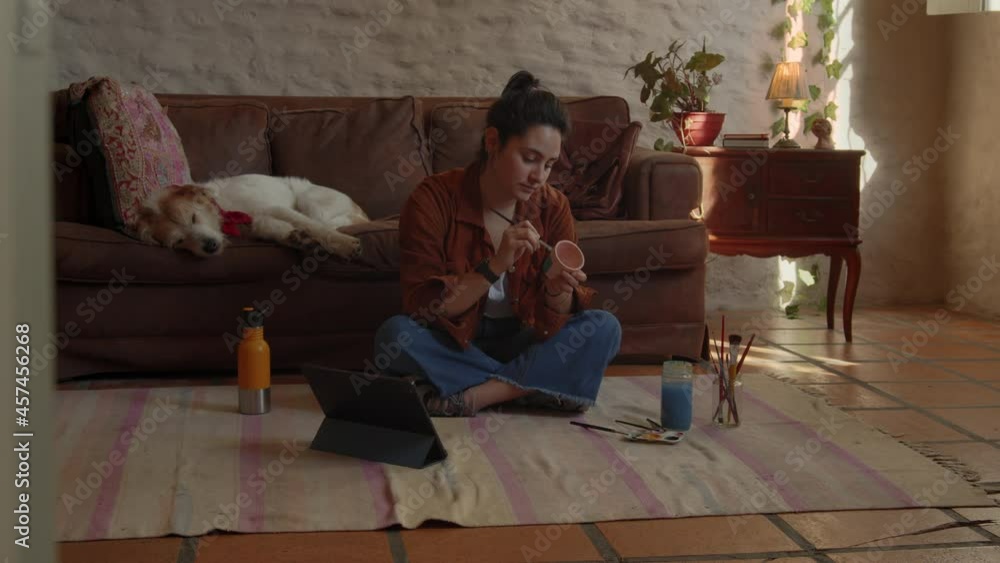 Woman painting flowerpot sitting on a carpet and checking her hand watch, discovery shot Woman painting flowerpot sitting on a carpet and checking her hand watch, discovery shot