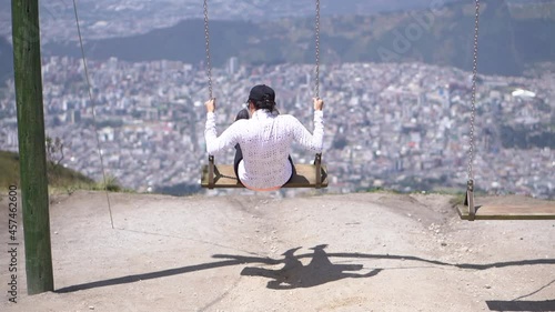 A woman wearing sports clothes is swinging on a swing on top of a mountain near the city of Quito, Ecuador. Panoramic view of the city of Quito.