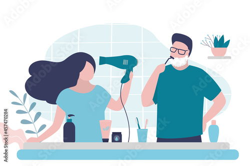Girl dries wet hair with hairdryer. Man shaves stubble with razor and shaving foam. Couple doing morning routine together