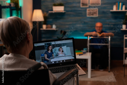 Aged woman with handicap using video call with laptop at home. Senior person talking to daughter and child while sitting in wheelchair in living room with online remote conference © DC Studio