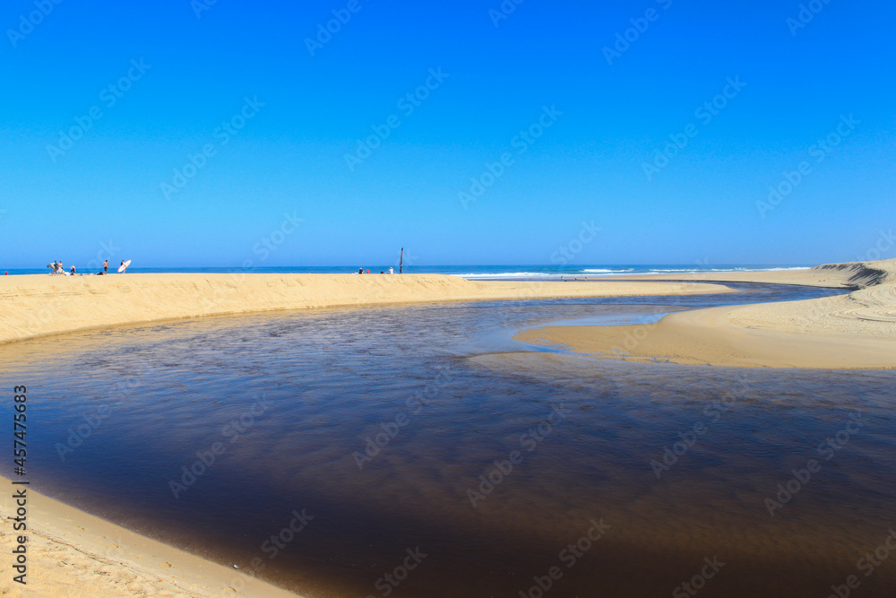Obraz premium Le courant d'Huchet serpente au milieu du sable de la plage de Moliets dans les Landes pour finir par se jeter dans l'océan. Le ciel est bleu, il fait beau, c'est l'été