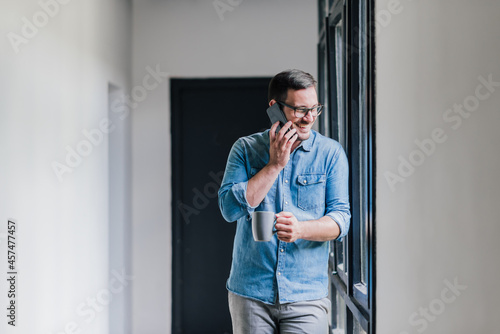 Confident smiling young successful cheerful casual entrepreneur businessman on coffee break talking on the phone making call satisfied drinking coffee leaning on big bright window