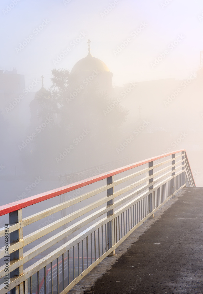 Fototapeta premium Red railings of a pedestrian bridge on the background of a temple in the fog in Novosibirsk