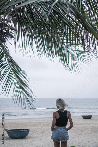Photo of pretty caucasian young woman with blond hair, wearing denim shorts and short top. Standing on the beach. Backside view. Palm branch.