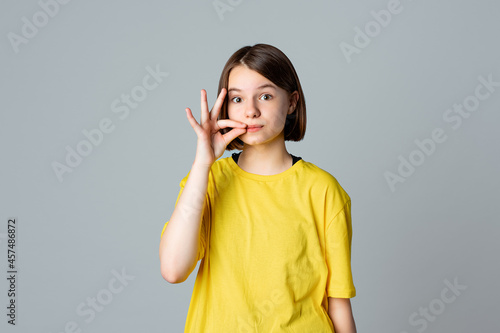 Portrait of a pretty teen girl showing zip gesture as if shutting mouth on key, standing over light grey background. You must be silent. Ban. Taboo. Censorship and harassment of freedom of speech photo