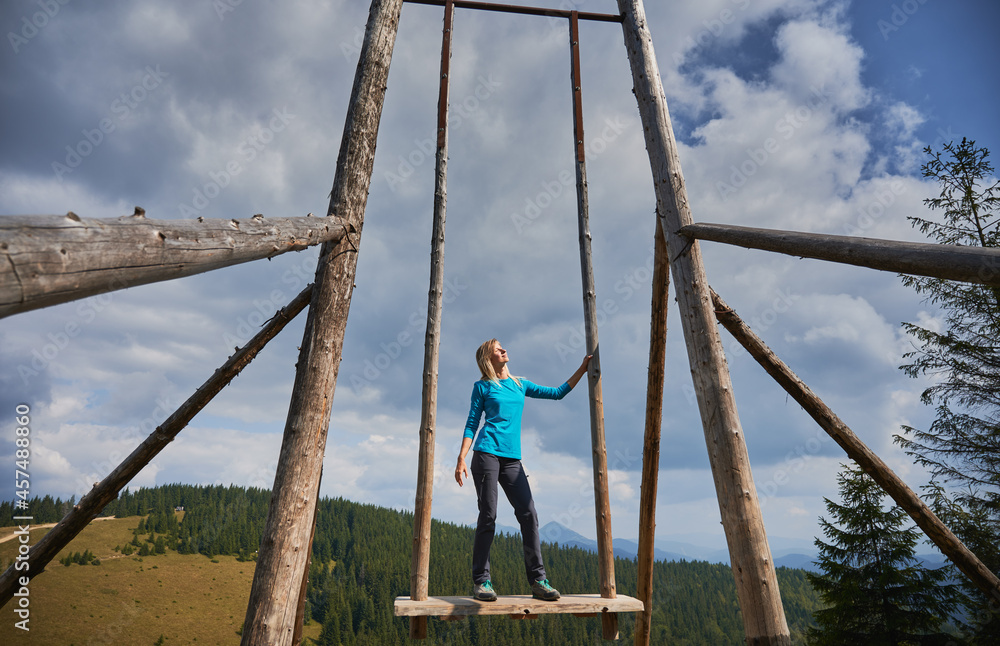 Against the backdrop of cloudy sky and mountain hills, female hiker relaxing, standing on seat of large wooden swing, basking in warm sun with closed eyes. Concept of travelling, freedom and nature. Against the backdrop of cloudy sky and mountain hills, female hiker relaxing, standing on seat of large wooden swing, basking in warm sun with closed eyes. Concept of travelling, freedom and nature.