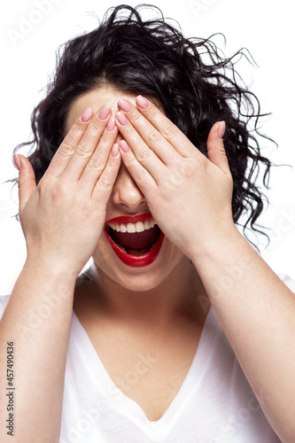 Laughing young woman covered her eyes with her hands. Beautiful brunette with red lips and curly hair. Positiveness and joy. Close-up. Vertical. White background.