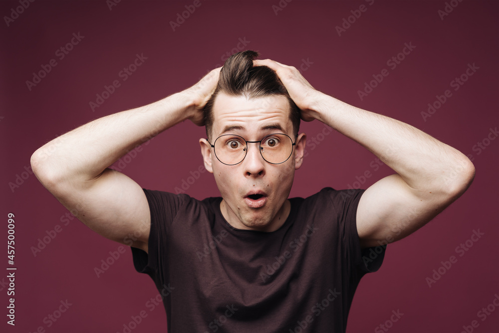 Surprised male, keeps mouth opened, being stupefied as advertises something, wears glasses, brown t shirt, holding hands in hair, over crimson background. Omg concept Surprised male, keeps mouth opened, being stupefied as advertises something, wears glasses, brown t shirt, holding hands in hair, over crimson background. Omg concept
