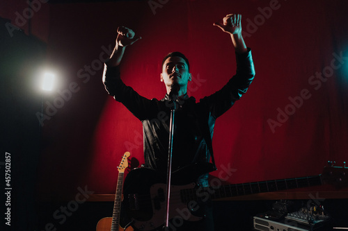 Rock band vocalist with the guitar singing to microphone with the hands raised up in lights on red background