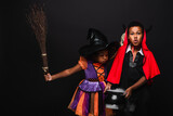 african american girl in halloween costume holding broom and blowing on cauldron with potion near brother isolated on black