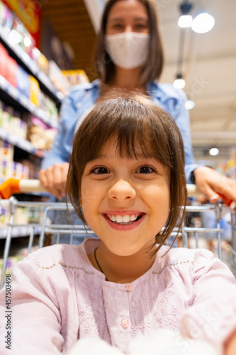 Happy girl sitting in the grocery cart making selfie with her mother wearing medical mask being in the store among the shelves with the goods.