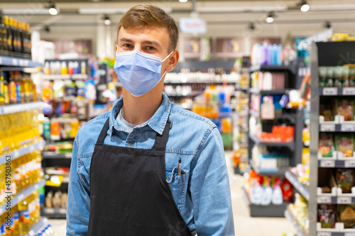 Store worker man wearing denim shirt with long sleeve having a pen in the chest pocket and apron black color posing in a face mask standing in the middle of the store market on pandemic time.