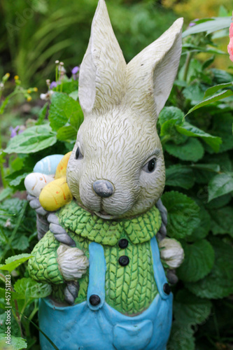 garden figure Easter bunny in blue jumpsuit with a backpack of colored eggs behind the back. 
