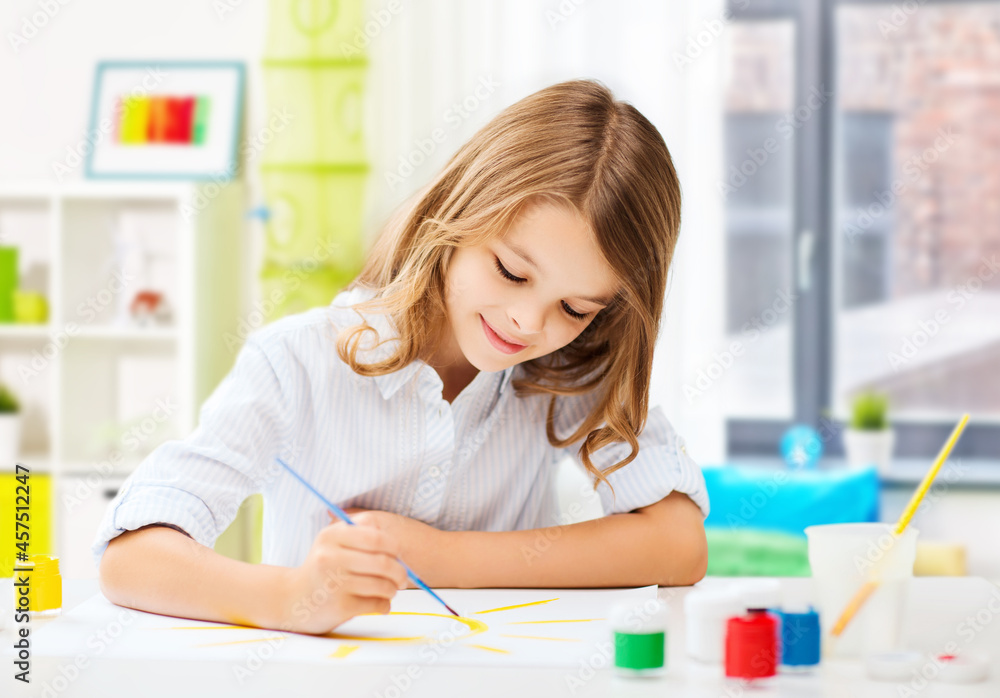 hobby, art and childhood concept - happy smiling girl with brush and colors drawing picture over home room background hobby, art and childhood concept - happy smiling girl with brush and colors drawing picture over home room background