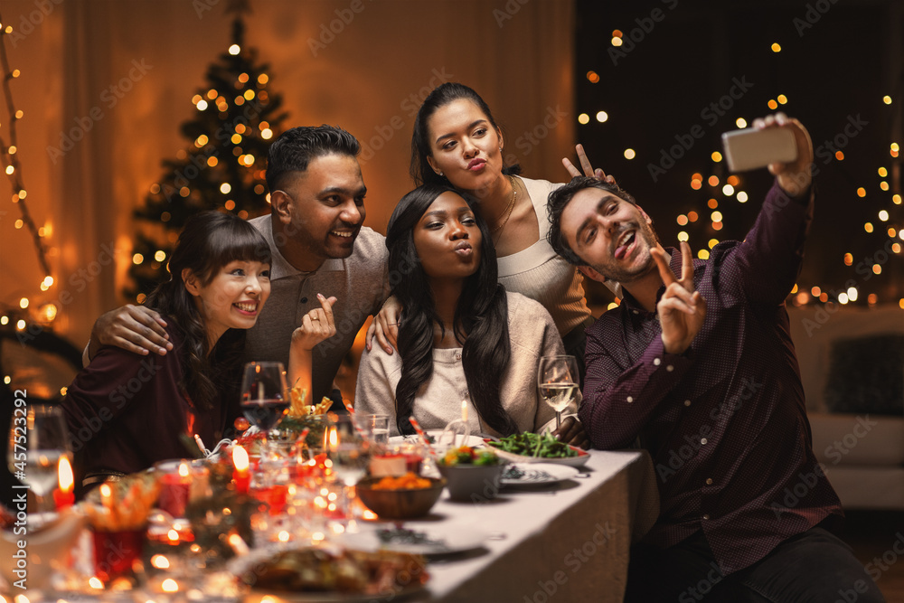 holidays, party and celebration concept - multiethnic group of happy friends having christmas dinner at home and taking selfie with smartphone