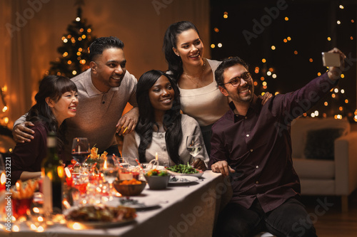 holidays  party and celebration concept - multiethnic group of happy friends having christmas dinner at home and taking selfie with smartphone