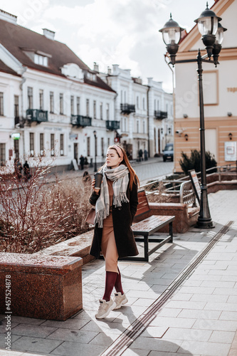 Stylish young beautiful girl with ginger hair walks around the city in autumn clothes with a coat and a scarf. Outfit for cold weather © Daria Lukoiko