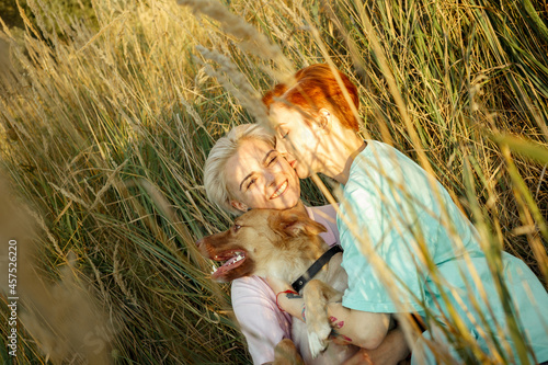 Joyful young lesbian couple with playful red dog has fun lying together in high field grass at sunset light in summer evening