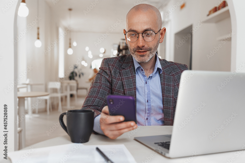 Adult bald man writes a message on a mobile phone while working at a laptop in a cafe Adult bald man writes a message on a mobile phone while working at a laptop in a cafe