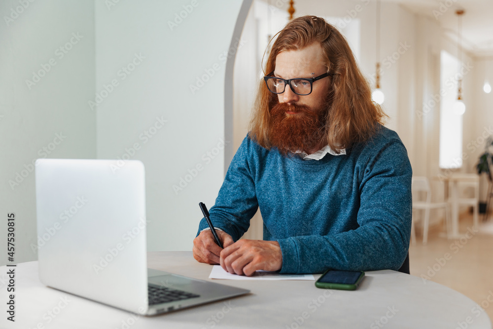 A man works at a laptop in a cafe and writes a note on paper A man works at a laptop in a cafe and writes a note on paper