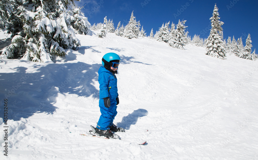 toddler boy in helmet, glasses and blue winter coveralls on skis in beautiful snow-capped mountains. Winter active entertainment for children. Skiing lesson at an alpine school. It's cold, sunny